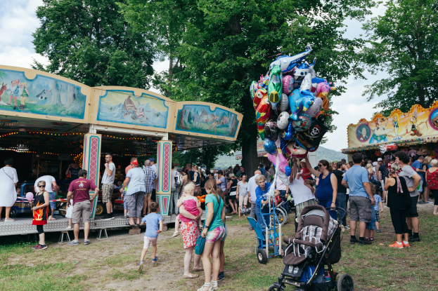 Eine Menschenmenge, einige mit Kinderwagen, versammelte sich um ein bunt beleuchtetes Karussell auf einem Volksfest, mit Tieren und einem bewölktem Himmel im Hintergrund.