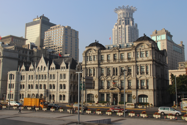 Eine belebte Stadtstraße mit einem prominenten zentralen Gebäude, umgeben von anderen Gebäuden, Fahrzeugen, Fußgängern, Bäumen und Straßeninfrastruktur unter einem klaren blauen Himmel.