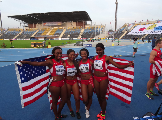Vier Frauen in Sportkleidung stehen zusammen auf einer Laufbahn, jede hält eine amerikanische Flagge und lächelt, mit einem Stadion und Zuschauern im Hintergrund.