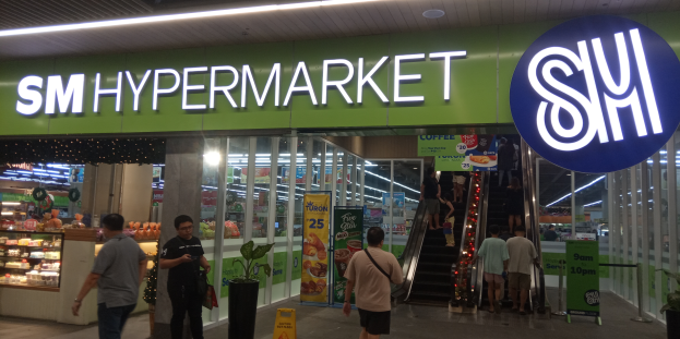 Eine Gruppe von Menschen, darunter Männer und Frauen, geht vor einem hell erleuchteten Geschäft mit einer Tafel, auf der 'SM Hypermarket' steht, vorbei, mit sichtbaren Schildern, Topfpflanzen und anderen Gegenständen im Inneren.