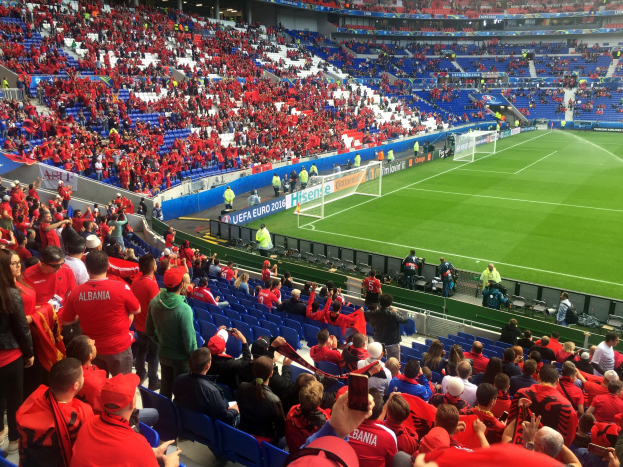 Eine große Menschenmenge in einem Stadion schaut einem Fußballspiel zu, mit Menschen auf Sitzplätzen und Stehplätzen, Bannern, einem Torpfosten und Deckenbeleuchtung.