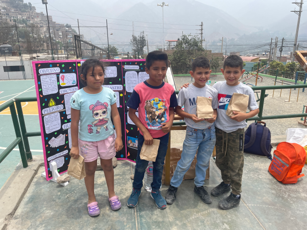 Eine Gruppe von Kindern steht auf einem Basketballplatz, jedes hält einen Papiertüte, mit Tüten und Gegenständen rechts und einer Tafel mit Text links, umgeben von Zaun, Pfosten, Straßenlaternen, Bäumen, Gebäuden, Hügeln und dem Himmel.