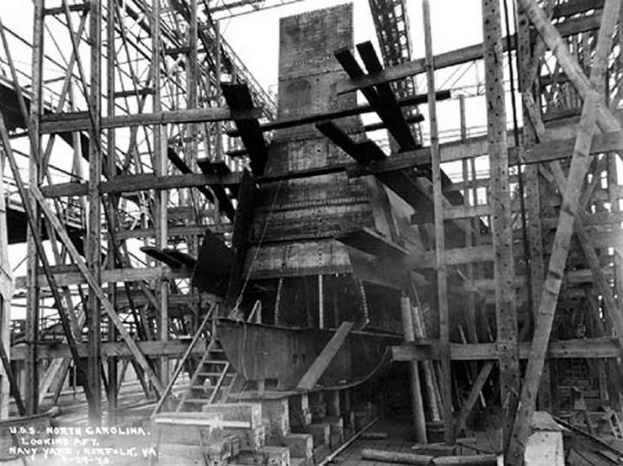 Schwarzes und weißes Foto eines Schiffs in einem Schiffbauwerk mit hölzernen Pfählen und Treppen im Vordergrund und Himmel im Hintergrund, mit dem Text "Schiffsbewaffnung" in der linken unteren Ecke.