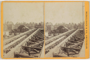 Ein altes Schwarz-Weiß-Foto einer Bahnschiene mit Gebäuden im Hintergrund, Text auf der rechten Seite mit der Aufschrift "Merkelbach & Co, Co. Amsterdam" und verstreute hölzerne Gegenstände im Vordergrund.