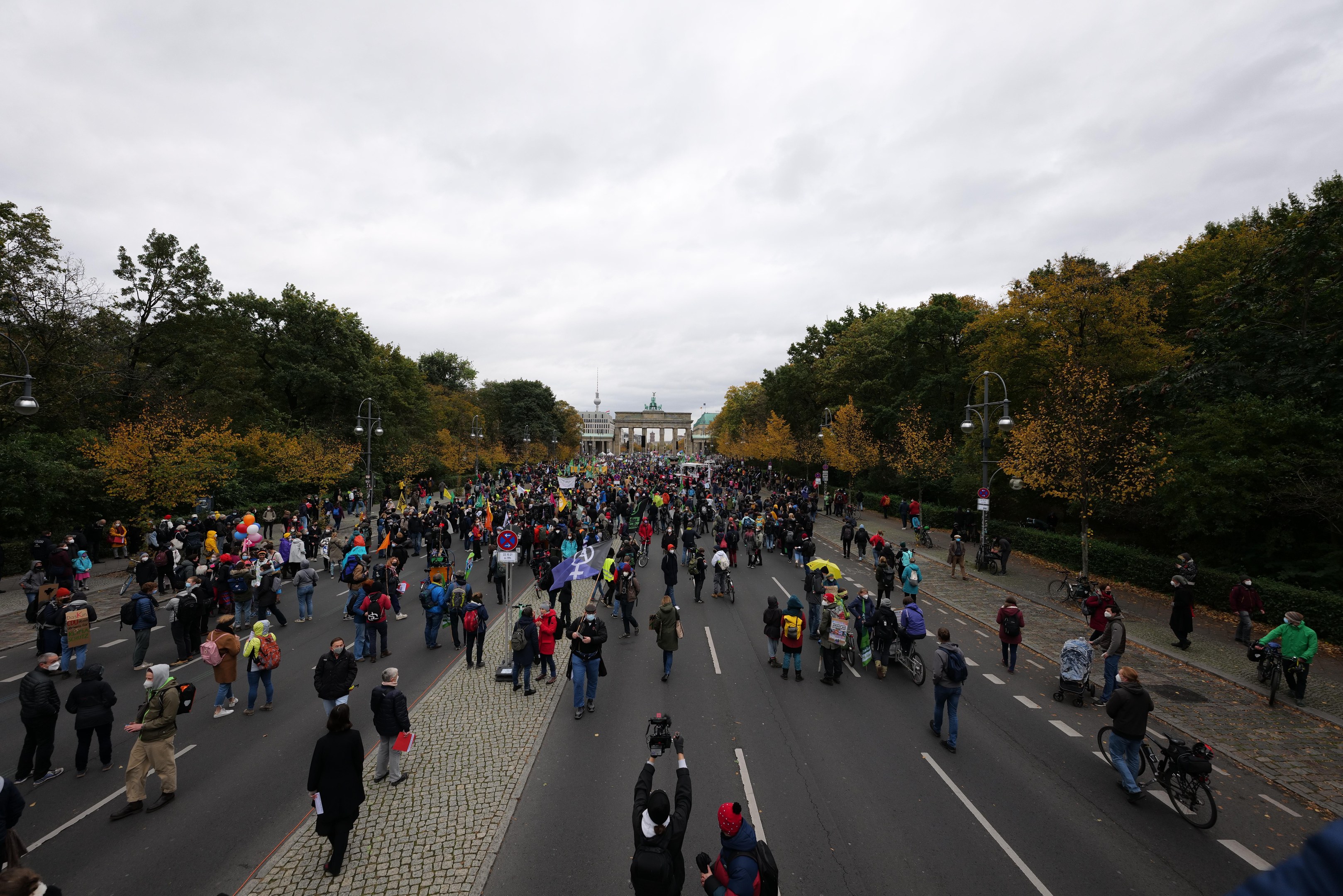 Eine große Gruppe von Menschen marschiert auf einer von Bäumen gesäumten Straße mit Laternenmasten, die Kameras halten, mit einem Gebäude und einem klaren Himmel im Hintergrund, was auf eine Demonstration in Berlin hinweist.