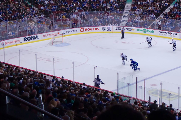 Vier Männer spielen Hockey auf einem Eisstadion, mit zwei stehenden nahe am Netz und Zaun, Zuschauern auf Stühlen im Vorder- und Hintergrund und einer Treppe im Hintergrund.