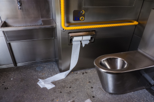 Eine Edelstahltoilette mit einer Rolle Toilettenpapier daneben, ein Waschbecken mit einem Wasserhahn und verschiedene Gegenstände, die auf dem Boden verstreut sind.