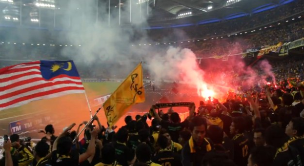 Große Menschenmenge in einem Stadion mit brennenden Fackeln in der Luft, einige halten Fahnen und eine Person hält eine Kamera, Rauch im Hintergrund, Schilder mit der Aufschrift "Malaysia vs Malaysia Live Streaming", Stadionbeleuchtung und -strukturen sind sichtbar.