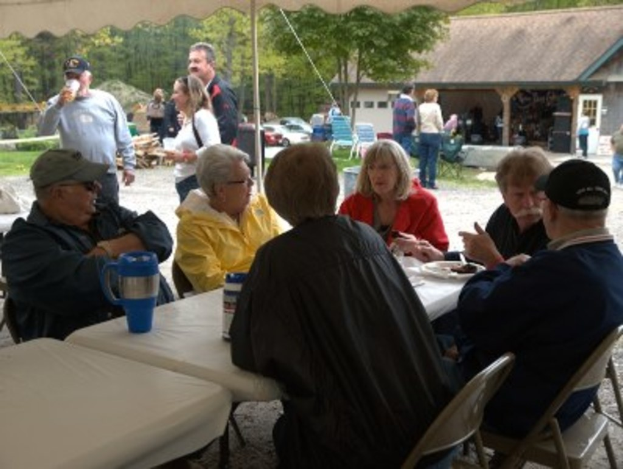 Ein Cafe-Innenraum mit zwei weißen Tischen, sechs Personen, die darum sitzen, eine Flasche und Essen auf dem Tisch, eine Gruppe von Menschen dahinter, zwei Personen rechts, ein Haus davor und ein Hintergrund aus Bäumen, Autos und Gras.