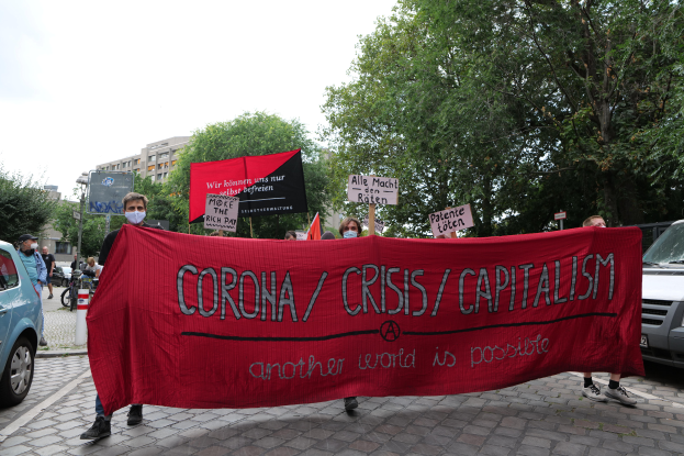 Eine Gruppe von Menschen marschiert mit einer roten Fahne, auf der "Corona-Krise/Kapitalismus" steht, eine Straße entlang. Parkende Fahrzeuge und Gebäude, Bäume und ein klarer blauer Himmel sind im Hintergrund zu sehen.