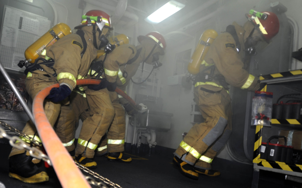 Feuerwehrleute in Schutzausrüstung arbeiten an einem Feuerwehrauto, mit Equipment-Racks und Hintergrund-Infrastruktur im Sichtfeld.
