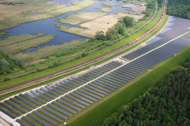 Luftaufnahme eines Solarparks mit Panelen in einem Feld, umgeben von Bäumen und Wasser, mit einem Zug auf einer nahen Schiene.