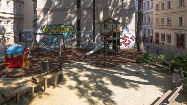 Ein Spielplatz mit Graffiti, umgeben von Gebäuden, Bäumen, Bänken, Pfählen und einem Mülleimer, mit Menschen und Fahrzeugen auf der Straße und dem Himmel im Hintergrund.