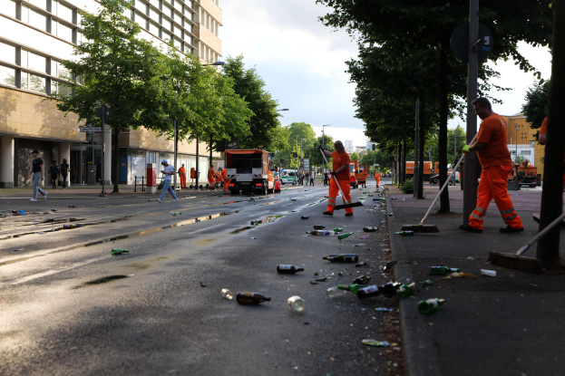 Eine Gruppe von Menschen in orangefarbenen Uniformen sammelt Müll am Straßenrand, mit Flaschen und anderen Gegenständen, die auf der Straße, an Bäumen, Pfählen, Brettern und Fahrzeugen in der Nähe sowie Gebäuden mit Fenstern und einem bewölkten Himmel im Hintergrund verstreut sind.