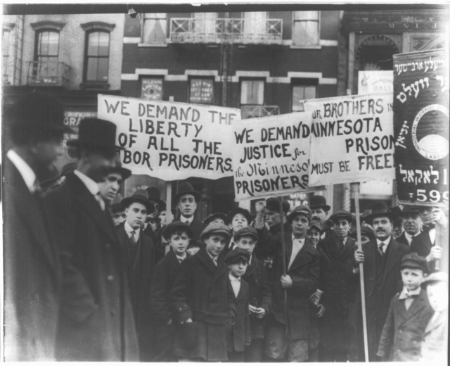 Schwarzes und weißes Foto einer Gruppe von Menschen in einer Reihe mit Schildern, auf denen "Wir fordern die Freiheit aller der Gerechtigkeit für die Gefangenen" steht, vor einem Gebäude mit Fenstern und einem Geländer.