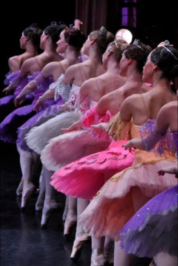 Eine Gruppe von Balletttänzerinnen in rosafarbenen Tutus sitzt auf einer Bühne, beleuchtet von einem Licht im Hintergrund, das sich in perfekter Harmonie bewegt.