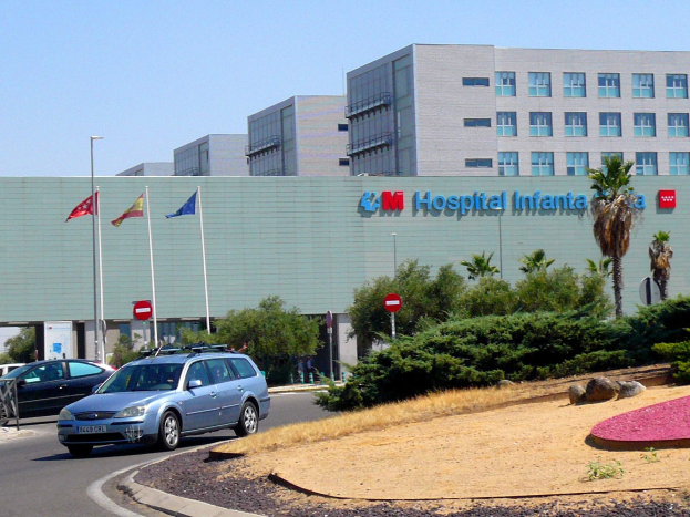 Krankenhaus mit parkenden Autos, umgeben von städtischen Elementen unter einem klaren blauen Himmel.