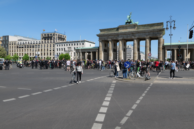 Eine große Gruppe von Menschen, die Fahrräder auf einer Straße vor dem Brandenburger Tor in Berlin, Deutschland fahren, mit Bäumen, Gebäuden, Laternenmasten und Verkehrszeichen an der Straße und einem Bogen mit Säulen und einer Statue im Hintergrund unter einem sichtbaren Himmel.