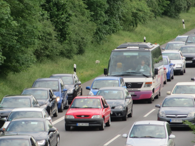 Ein Stau auf einer Autobahn mit vielen Autos und einem Lieferwagen, Menschen sind in den Fahrzeugen zu sehen, Bäume und Gras im Hintergrund.