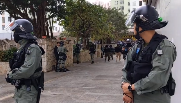 Eine Gruppe von Polizisten in Helmen und Schutzwesten steht auf einer Straße in Hong Kong, mit Bäumen, Gebäuden und einem klaren Himmel im Hintergrund.