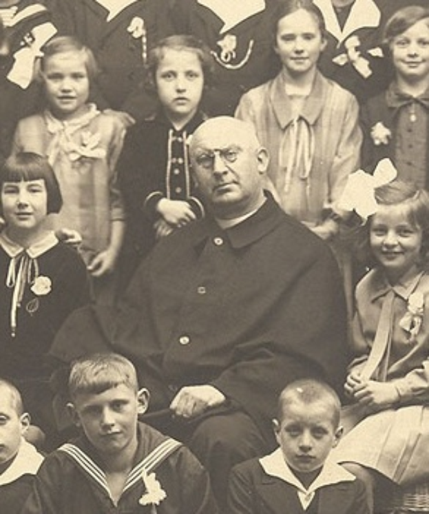 Ein altes Schwarz-Wei├č-Foto eines ernst aussehenden Mannes, der in der Mitte sitzt und von einer Gruppe von Kindern umgeben ist, die alle bewundernd zu ihm aufschauen.
