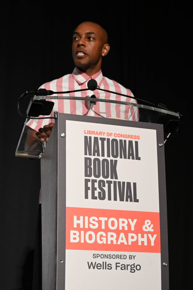 Ein Mann spricht an einem Podium mit einem Mikrofon vor einem Schild, auf dem "National Book Festival History & Biography" steht, bei dunkler Abendbeleuchtung.