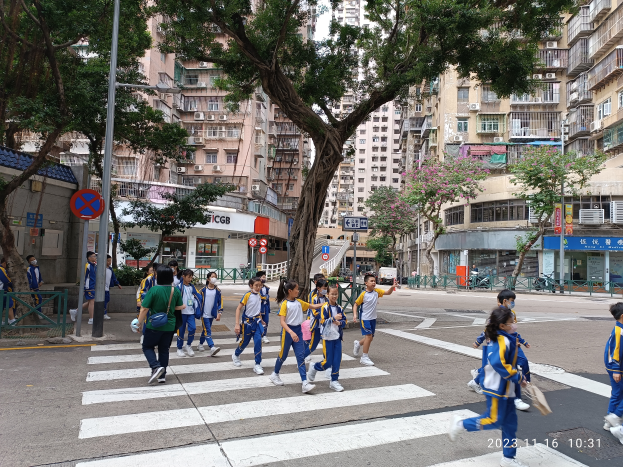 Eine Gruppe von Kindern in blauen und weißen Uniformen überquert eine Straße mit Bäumen, Laternen und Schildern, mit hohen Gebäuden und einem klaren Himmel im Hintergrund.
