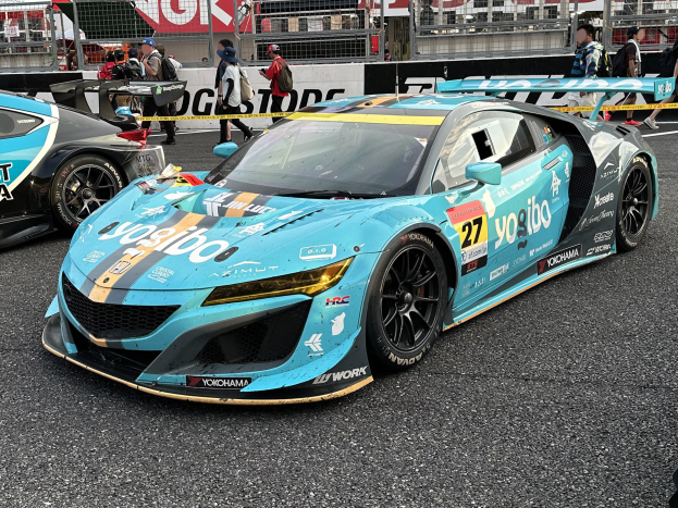 Gruppe von Rennwagen auf einer Rennstrecke mit Menschen in Mützen und Taschen im Hintergrund, umgeben von Schildern, Geländern, Pfosten und einem fernen Gebäude, mit "Yokohama Acura NSX GT3"-Text unten.
