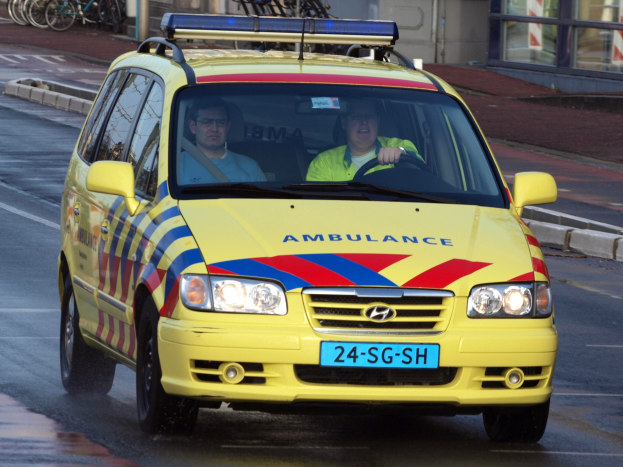 Ein gelber Krankenwagen mit zwei sichtbaren Insassen fährt an parkenden Fahrrädern auf einem Fußweg vorbei, mit einem Glasfenstergebäude im Hintergrund.