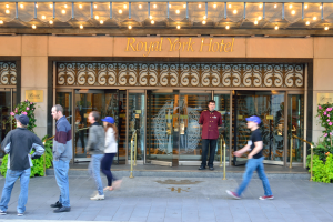 Eine Gruppe von Menschen, die vor dem Royal York Hotel in New York City gehen, mit Text an der Wand, Glastüren, Lampen und von blühenden Pflanzen umgeben.