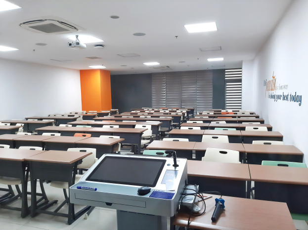 Ein Klassenzimmer mit Tischen und Stühlen in Reihen, einem deckengehängten Projektor und Gegenständen auf den Tischen, mit Text an der rechten Wand, der darauf hinweist, dass es sich um eine Privatschule in Singapur handelt.