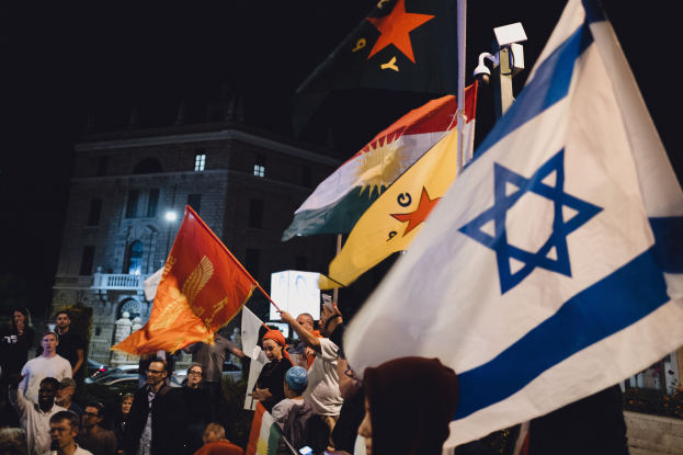 Eine Gruppe von Menschen hält Flaggen vor einem Gebäude mit Fenstern und einer Straßenlaterne, mit Pflanzen und Fahrzeugen im Hintergrund, unter einem dunklen Himmel während einer anti-israelischen Demonstration.