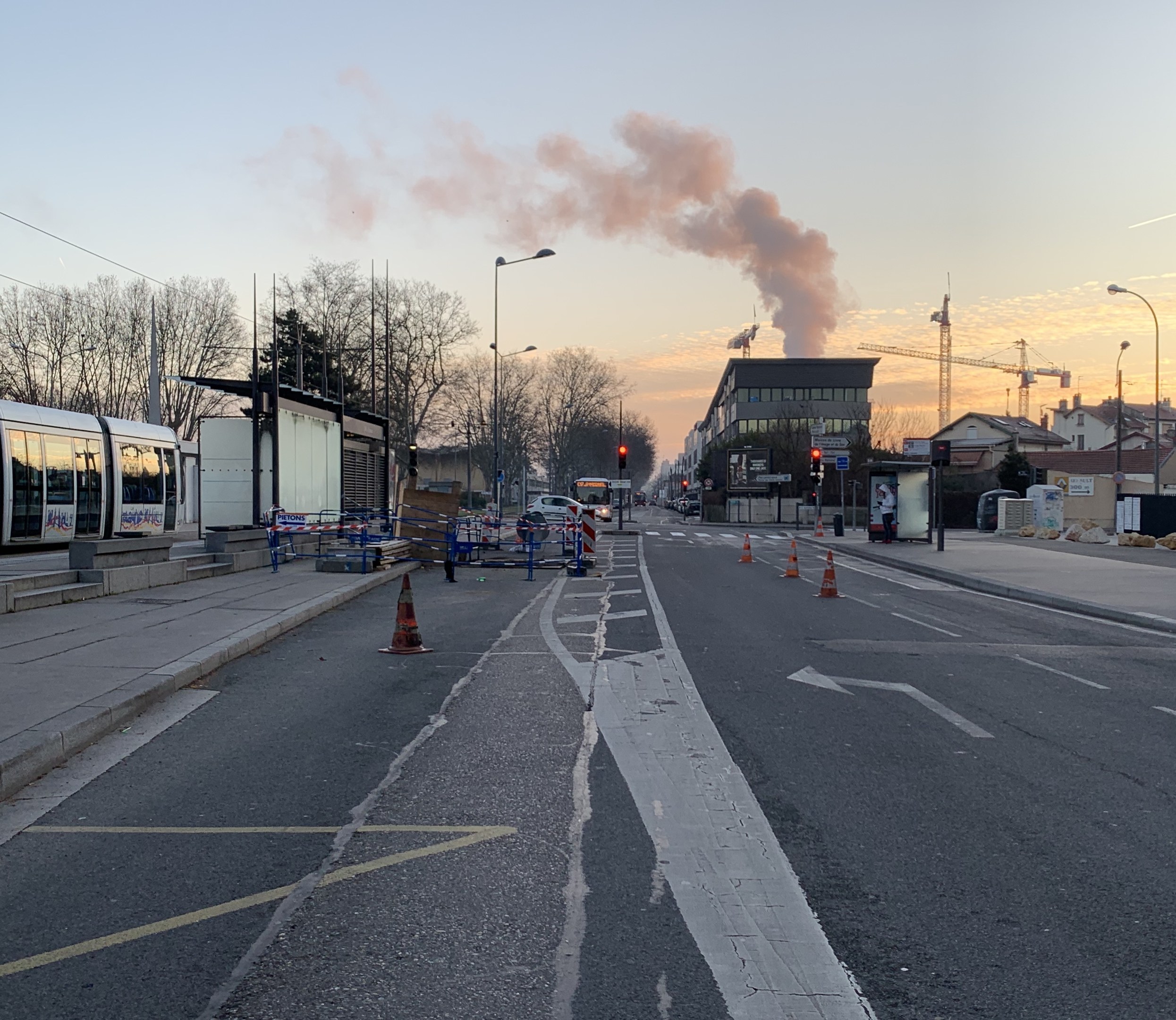 Strassenszene mit Verkehrskegeln, einem Bus, Laternen, Bäumen, Gebäuden, Kränen und Rauch, der aus einem entfernten Gebäude aufsteigt.
