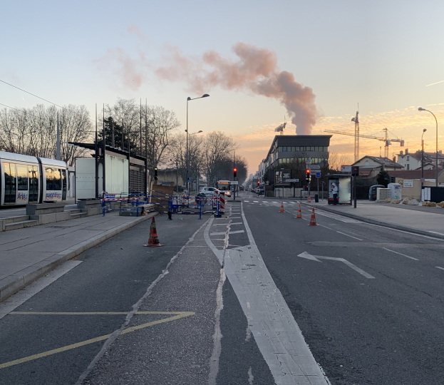 Strassenszene mit Verkehrskegeln, einem Bus, Laternen, Bäumen, Gebäuden, Kränen und Rauch, der aus einem entfernten Gebäude aufsteigt.
