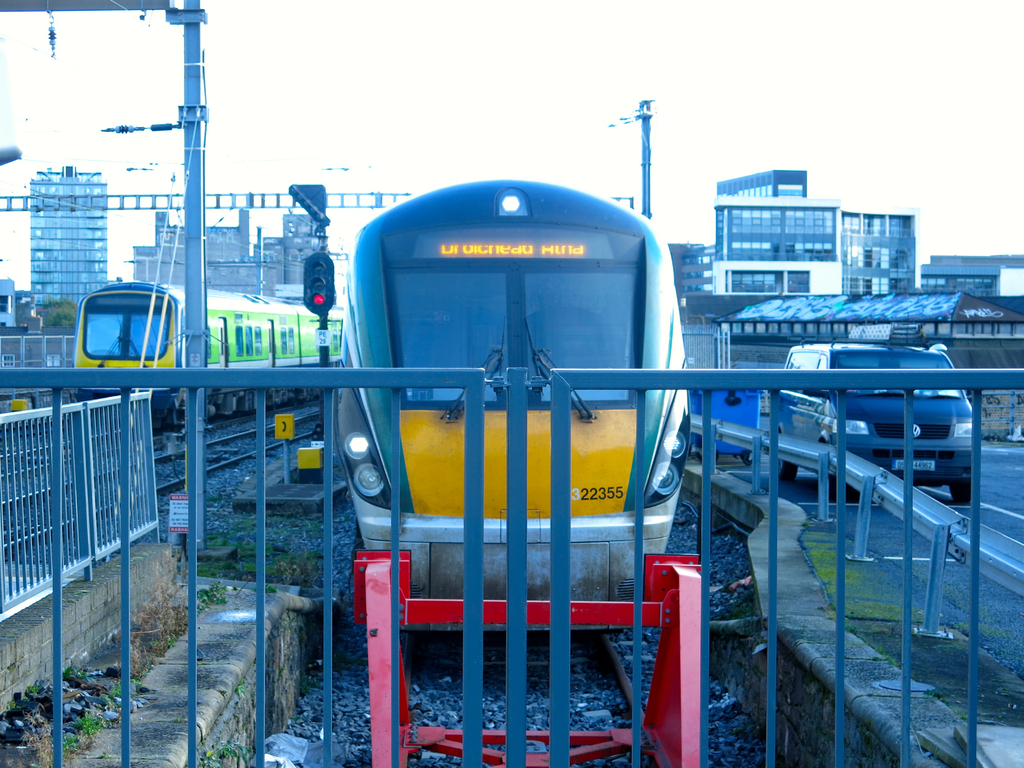 Zwei Züge befinden sich auf der Schiene mit einem Zaun im Vordergrund, Pfosten auf dem Land, einer mit einer Signalleuchte, ein Auto auf der Straße rechts, Gebäude im Hintergrund und der Himmel oben.