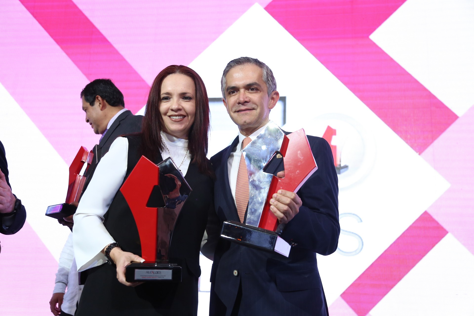 Zwei Menschen auf einer Bühne, die lächeln und Trophäen halten, mit einem Bildschirm im Hintergrund.