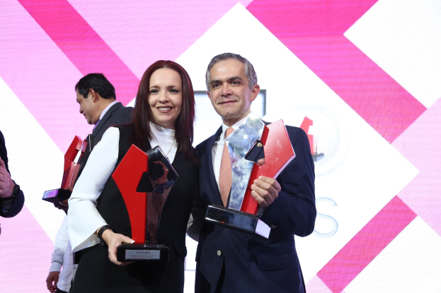 Zwei Menschen auf einer Bühne, die lächeln und Trophäen halten, mit einem Bildschirm im Hintergrund.