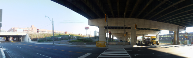 Ein Lastwagen fährt unter einer Brücke auf einer Stadtstraße mit Gebäuden, Straßeninfrastruktur, Fahrzeugen, Grünanlagen und einem klaren Himmel.