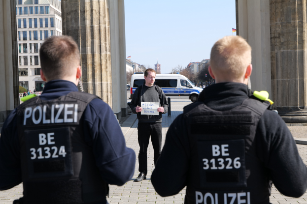 Drei Polizeibeamte stehen vor einem Gebäude, wobei ein Mann in der Mitte ein Papier hält, flankiert von Säulen, während im Hintergrund Fahrzeuge, Menschen, Bäume und Gebäude unter dem Himmel zu sehen sind.