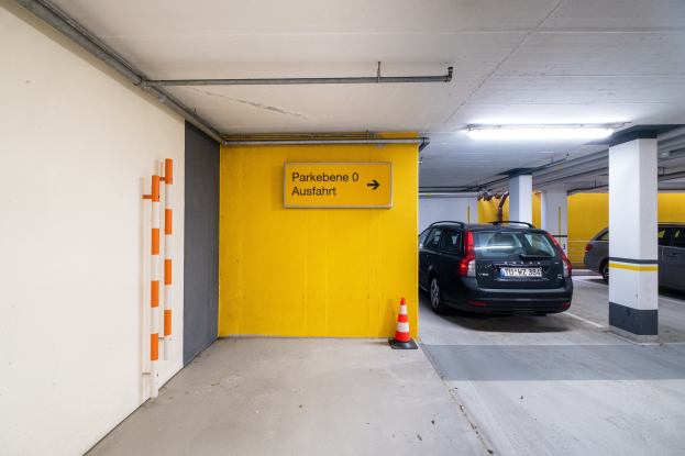 Eine Tiefgarage mit zwei geparkten Autos, einem Verkehrskegel auf dem Boden, einer Wandtafel mit Text, Deckenleuchten und einem sichtbaren Rad.