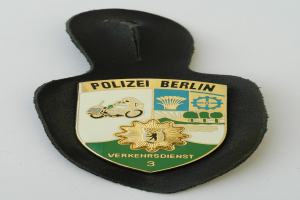 Ein schildförmiges Polizeisiegel mit 'Polizei Berlin' in schwarzer Schrift auf einem blauen Hintergrund mit einem weißen Stern und Rahmen, das auf einer Oberfläche liegt.