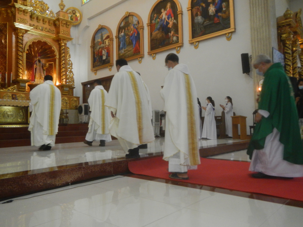 Eine Gruppe von Priestern geht einen roten Teppich in einer Kirche entlang, während Menschen vor ihnen auf dem Boden und auf der rechten Seite stehen und Fotorahmen an der Wand im Hintergrund zu sehen sind.