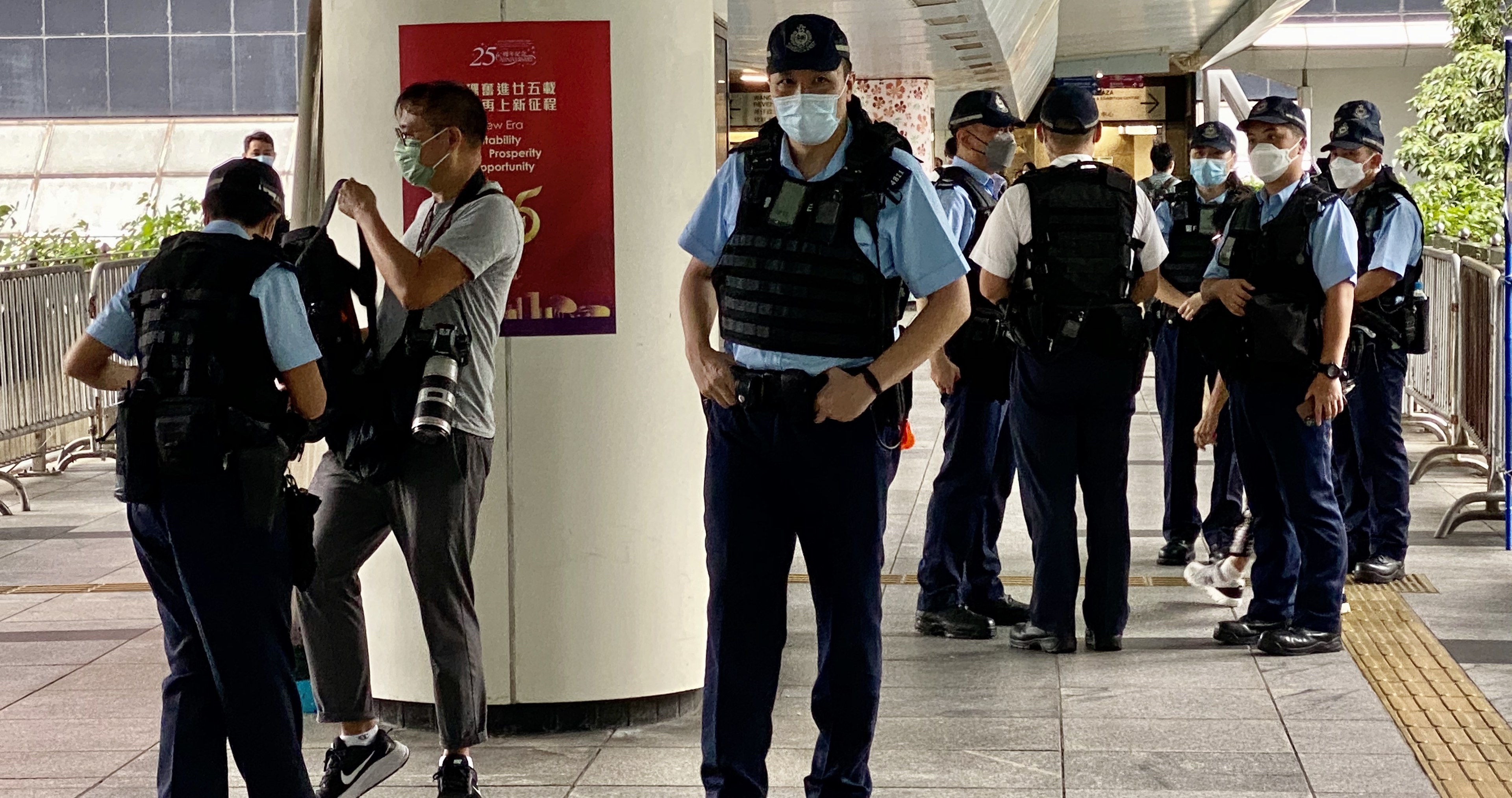 Polizeibeamte in Uniform und mit Mund-Nasen-Schutz stehen vor einem Gebäude, einige halten Kameras, mit einem Banner, Geländern, Pflanzen, Bäumen und Deckenleuchten im Hintergrund.