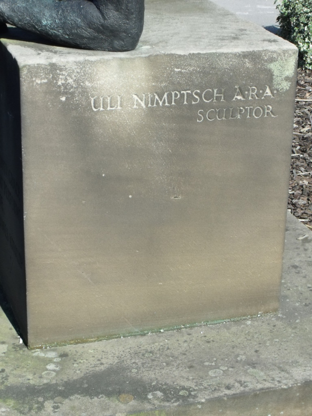 Ein Stein mit Text im Vordergrund, möglicherweise eine Statue oben, trockene Zweige, Erde und Pflanzen rechts, mit einer Straße im Hintergrund.