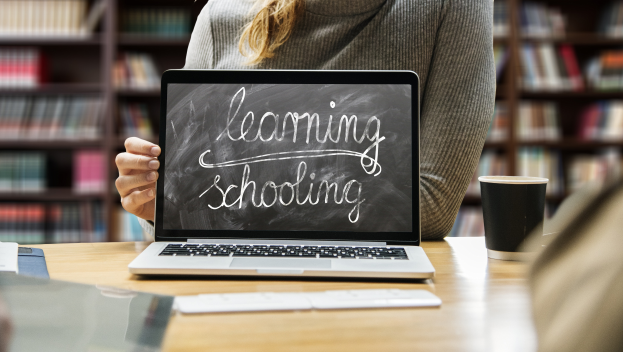 Eine Frau sitzt an einem Tisch mit einem Laptop, auf dem "Lernende Schule" angezeigt wird, umgeben von Papieren, einem Becher und anderen Gegenständen, mit Bücherregalen im Hintergrund.