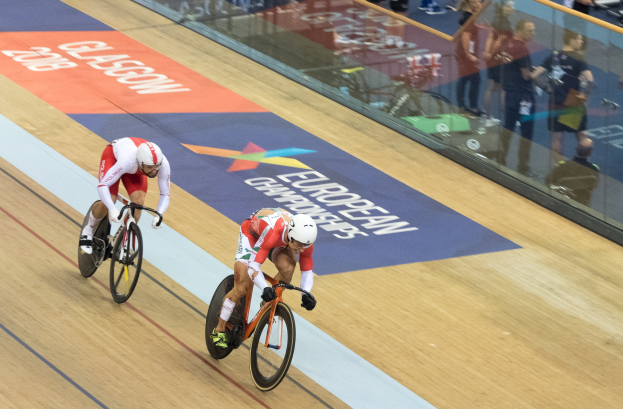 Zwei Männer mit Helmen fahren auf einer Indoor-Bahn vor einem Publikum, mit einer Glaswand, auf der 'Europameisterschaften Glasgow 2018' steht und Zuschauern dahinter.