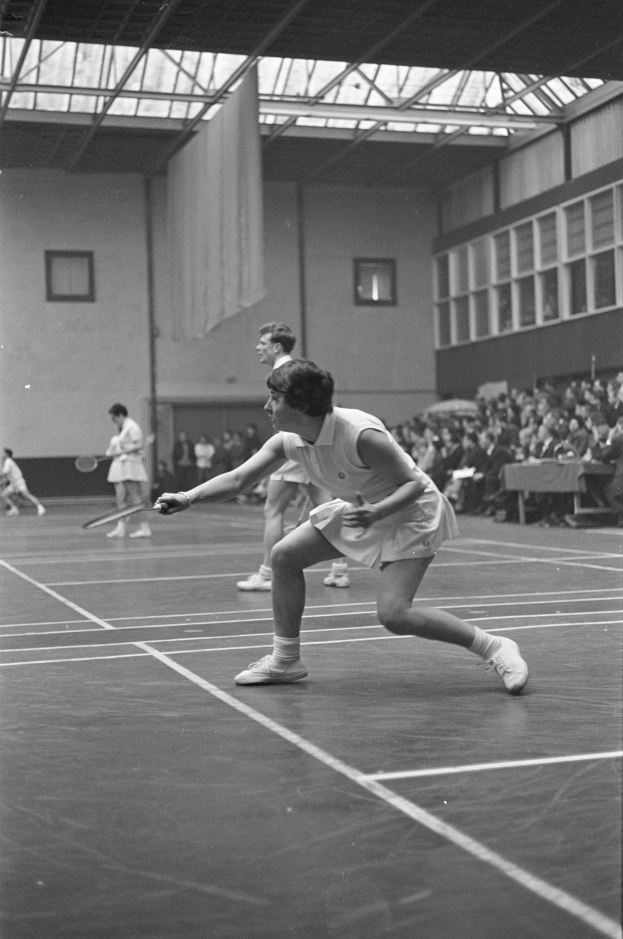 Schwarzes und weißes Foto von zwei Frauen, die Badminton auf einem Court spielen, jede hält einen Schläger in der Hand, mit Zuschauern rechts und einer Wand mit Fenstern im Hintergrund.
