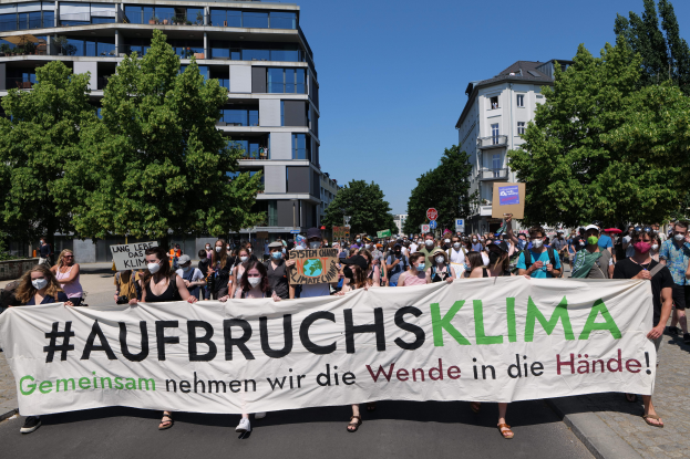 Eine Gruppe von Menschen mit Masken hält ein Schild mit der Aufschrift "Aufbruchsklima" vor einem Gebäude, umgeben von Bäumen unter einem klaren Himmel.