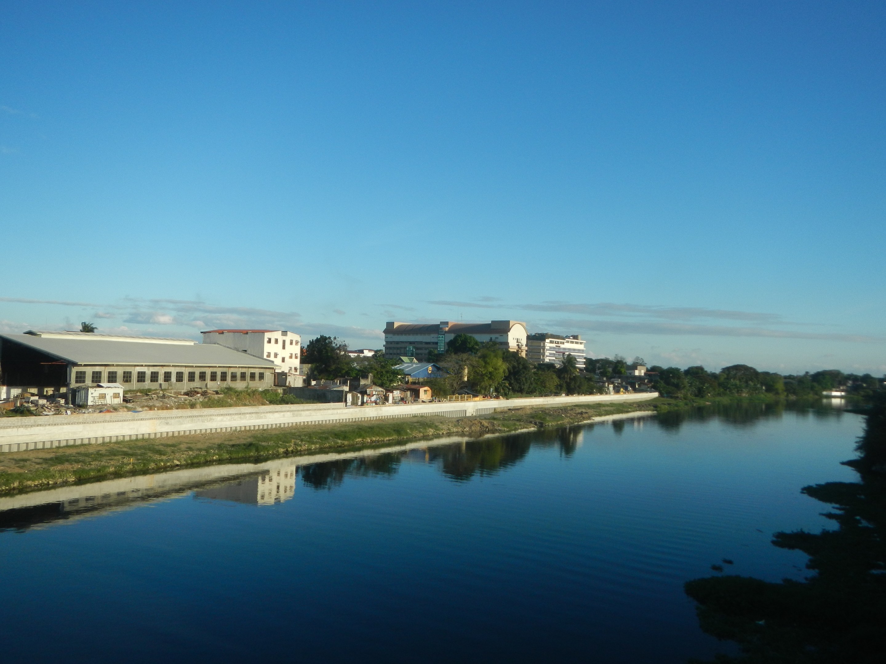 Ein ruhiger, klarer Fluss fließt durch eine grüne Wiese neben einer Stadt und spiegelt den Himmel mit Gebäuden, Bäumen und Wolken darüber wider.