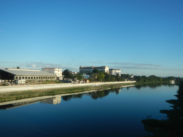 Ein ruhiger, klarer Fluss fließt durch eine grüne Wiese neben einer Stadt und spiegelt den Himmel mit Gebäuden, Bäumen und Wolken darüber wider.